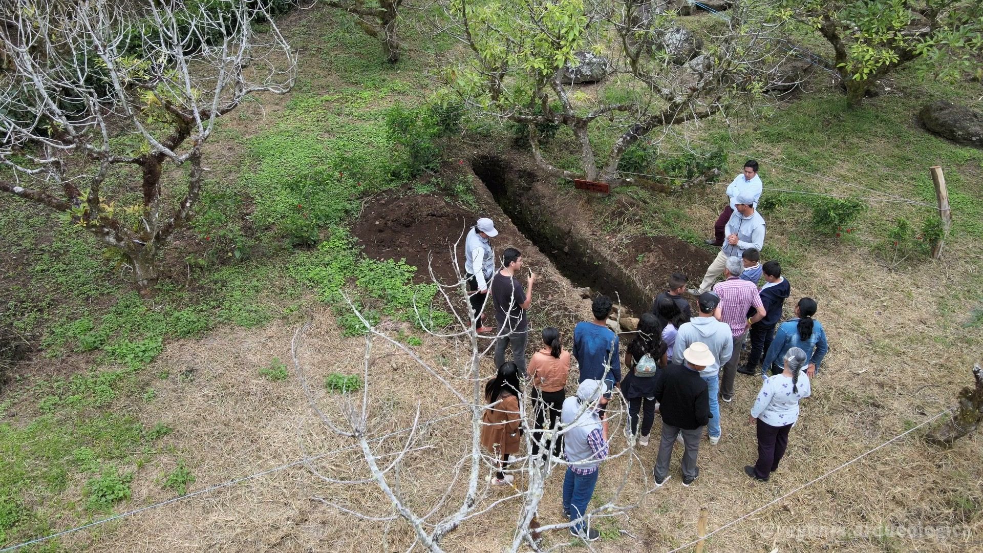 Un guía explicando una zanja de excavación abierta a un grupo de unos quince visitantes, con señalética y cuerdas de delimitación